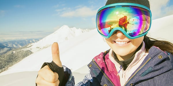 Femme avec lunettes de ski et vêtements d'hiver qui lève le pouce sur une montagne enneigée