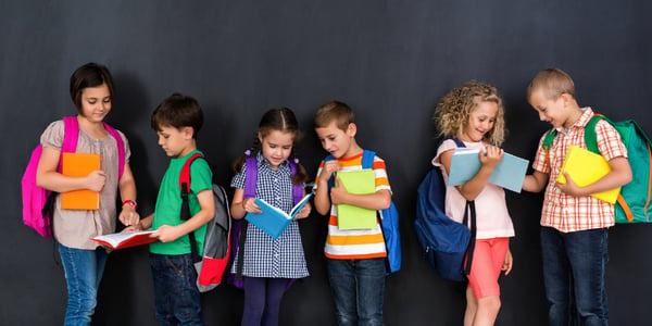 Enfants avec sac à dos et livres devant un tableau noir