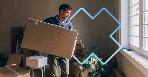 man carrying a large cardboard box during a move, inside a bright apartment filled with moving boxes