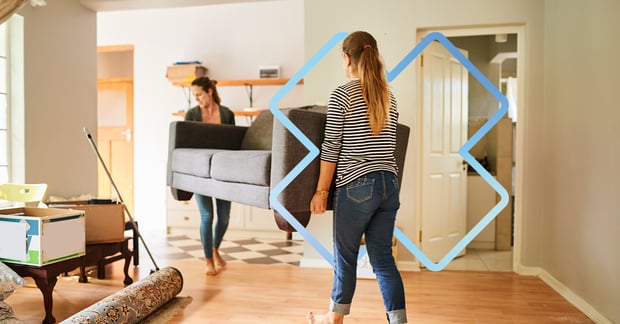 two women lifting a sofa during a home move, barefoot, surrounded by moving boxes and rolled-up rugs