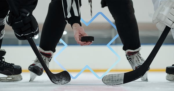 Deux joueurs de hockey sur glace s’apprêtent à disputer une mise au jeu, leurs bâtons croisés sur la glace. Un arbitre au centre tient le palet prêt à le lâcher. L’image est prise de près au niveau de la glace, mettant en valeur l’intensité du moment.