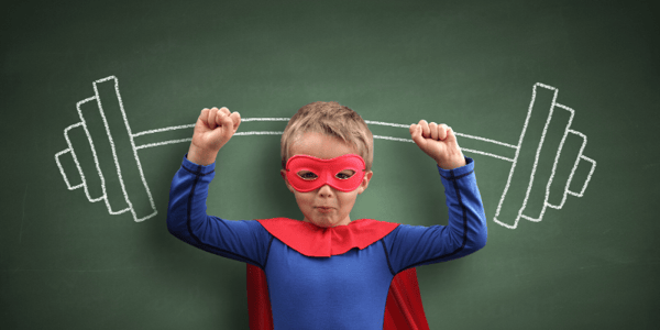 Enfant habillé en superhéro qui mime le soulèvement d'une haltère dessinée à la craie sur un tableau derrière lui